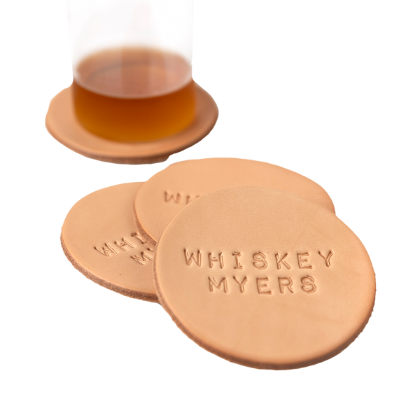 Leather coasters with ’Whiskey Myers’ embossed on them, alongside a glass of whiskey.