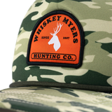 Camouflage-patterned baseball cap with an orange patch featuring a deer logo and ’Whiskey Myers Hunting Co.’ text.